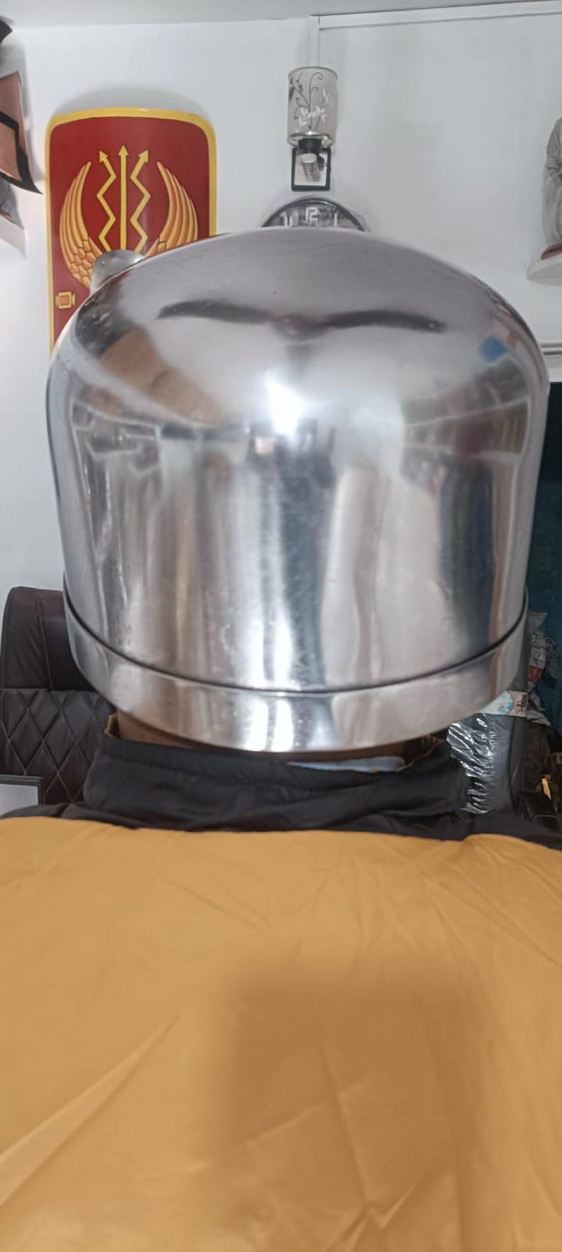 May include: A polished, silver-colored metal helmet with a smooth, rounded dome shape and a distinct horizontal seam encircling the lower portion. The surface is highly reflective, showing distorted light reflections across its curved exterior.