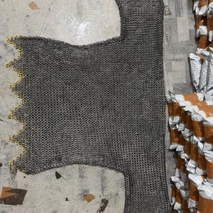 Puede incluir: Una cota de malla plateada con borde festoneado y ribete dorado. La prenda de estilo medieval está hecha de pequeños anillos de metal entrelazados. La camisa está tendida sobre una superficie gris.