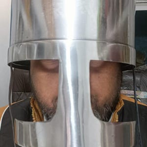 May include: A polished silver-colored metal helmet featuring a cylindrical top and a T-shaped face guard with a wide horizontal and vertical opening. The helmet has thin black straps attached to the sides and a smooth, reflective surface finish.