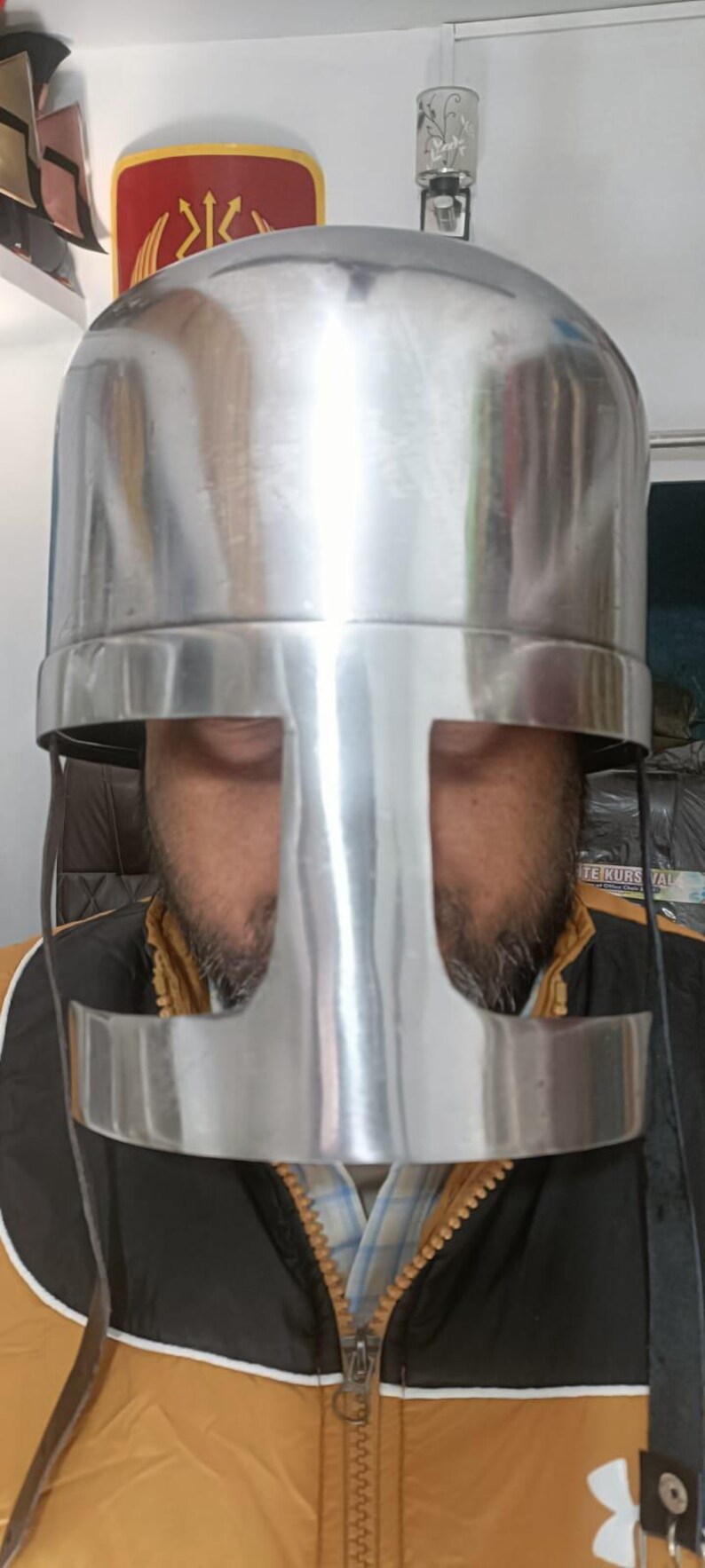 May include: A polished silver-colored metal helmet featuring a flat top and a T-shaped face opening. The helmet has a smooth, reflective surface and includes thin black straps hanging down from the sides of the visor area.