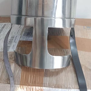 May include: A polished silver-colored metal helmet featuring a rounded dome top and a T-shaped face opening. The helmet includes two long black leather chin straps hanging down from the sides, with a metal buckle attached to one of the straps.