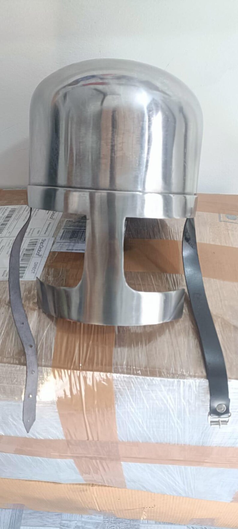 May include: A polished silver-colored metal helmet featuring a rounded dome top and a T-shaped face opening. The helmet includes two long black leather chin straps hanging down from the sides, with a metal buckle attached to one of the straps.
