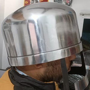May include: A polished silver-colored metal helmet with a rounded dome and a wide, curved face guard. The helmet features a smooth, reflective surface and a dark leather chin strap hanging down from the side, designed for historical reenactment.