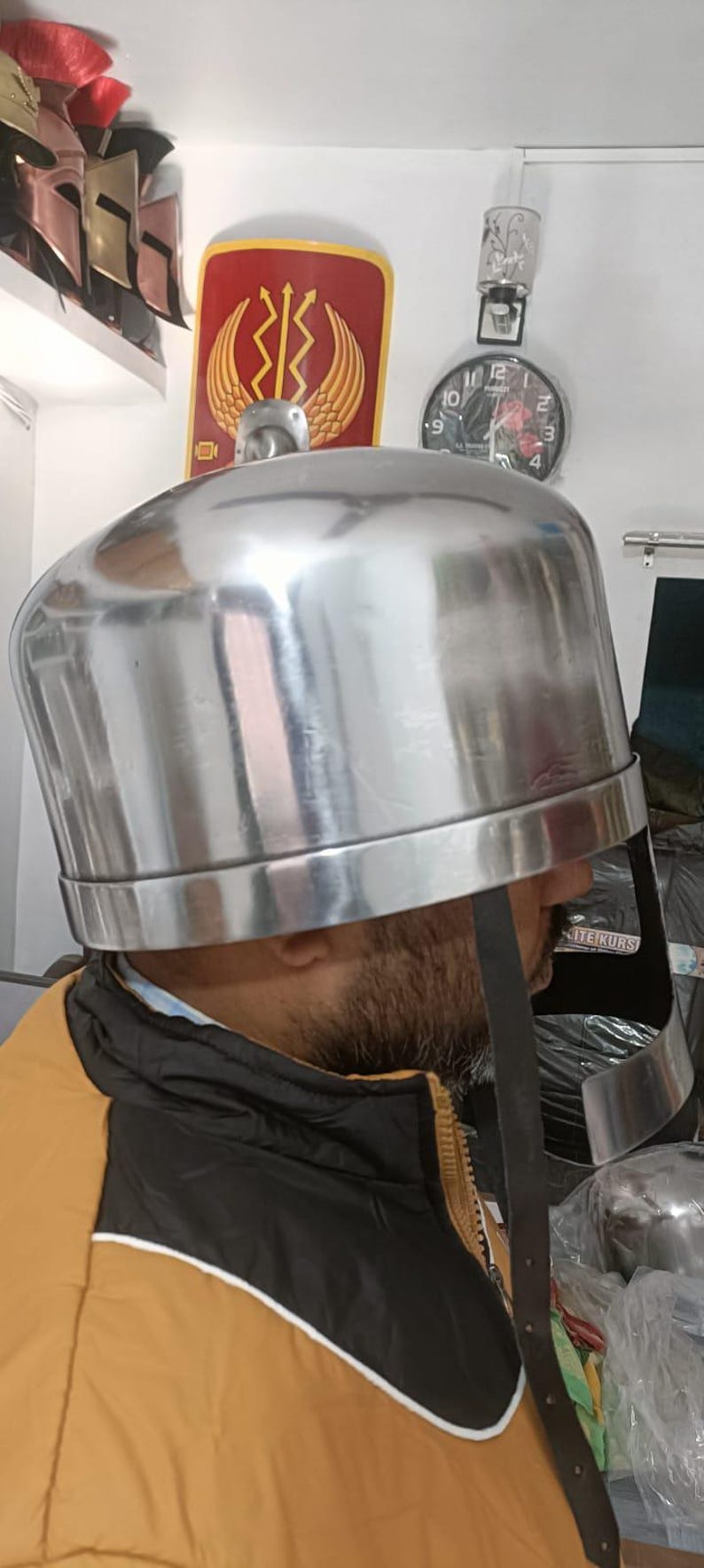 May include: A polished silver-colored metal helmet with a rounded dome and a wide, curved face guard. The helmet features a smooth, reflective surface and a dark leather chin strap hanging down from the side, designed for historical reenactment.