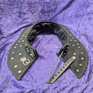 May include: A black, curved, wide collar accessory featuring multiple silver-toned metal studs arranged in rows across the surface. The collar includes a silver-toned metal buckle closure and a separate adjustable strap with pre-punched holes.