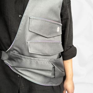 Può includere: Borsa a tracolla grigia con più tasche e finiture riflettenti. La borsa ha un design strutturato con tre tasche frontali e una chiusura con fibbia. La borsa si indossa sulla spalla e a tracolla.