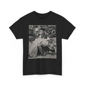 May include: A black short-sleeved t-shirt featuring a centered, rectangular, monochromatic graphic print. The image depicts a person with dark hair gesturing with one hand while interacting with a large, snarling feline creature in a natural setting.