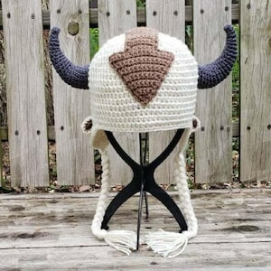 May include: A handmade, cream-colored crochet hat with dark blue horns and a brown arrow-shaped emblem. The hat features ear flaps and braided ties with tassels. The hat is displayed on a black stand against a wooden fence.