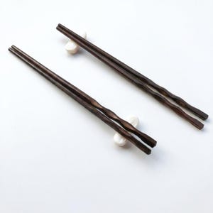 May include: Two pairs of dark brown wooden chopsticks with a wavy design, resting on small, white ceramic chopstick rests. The chopsticks are set against a clean, white background, highlighting their elegant form.