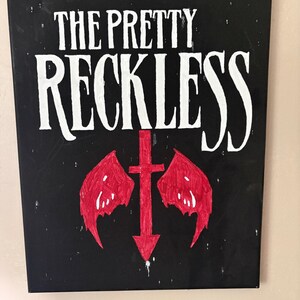 May include: A rectangular black canvas wall art piece featuring the white text THE PRETTY RECKLESS at the top. Below the text is a red painted graphic of a downward-pointing cross with two stylized red wings extending from the sides of the cross.