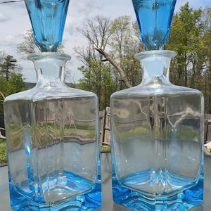Puede incluir: Dos decantadores de vidrio cuadrados con cuerpos transparentes y bases de color azul vibrante. Cada decantador cuenta con un tapón de vidrio azul facetado a juego que encaja en el cuello transparente. El diseño es simétrico con esquinas marcadas.
