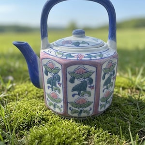 May include: A hexagonal ceramic teapot featuring a blue handle and spout, with a white body decorated with pink and green floral patterns. The teapot includes a matching lid with a small knob, and the exterior surface displays painted botanical motifs.