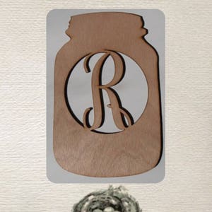 May include: Wooden cutout of a mason jar with a circle in the center that has the letter "R" cut out. The cutout is a light brown color.