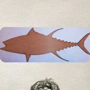 May include: A wooden cutout of a tuna fish on a light blue background. The tuna is brown and has a detailed outline.