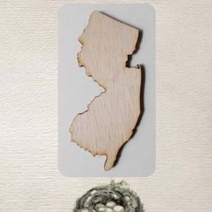 May include: A wooden cutout of the state of New Jersey. The cutout is light brown and has a natural wood grain.