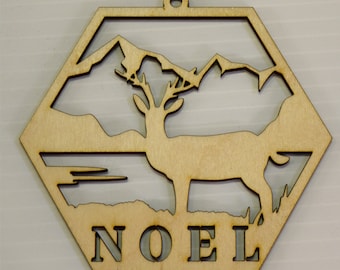 May include: Wooden Christmas ornament in the shape of a hexagon with a cut-out design of a deer standing in a mountain landscape. The word "NOEL" is cut out at the bottom of the ornament.