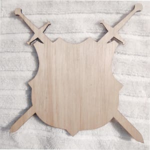 May include: A light-colored wooden shield cutout featuring a smooth, flat surface with a scalloped top edge. Two wooden sword cutouts are positioned behind the shield, crossing each other so that the hilts and blades extend from the sides.
