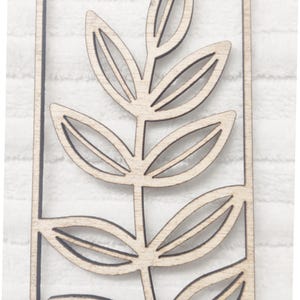 May include: A wooden cutout of a stylized branch with leaves. The cutout is framed in a square.