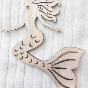 May include: A wooden cutout of a mermaid with a detailed tail and flowing hair. The mermaid is in a swimming pose with her tail extended behind her.