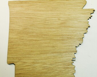 May include: A wooden cutout of the state of Arkansas. The cutout is made of light brown wood with a wood grain pattern.