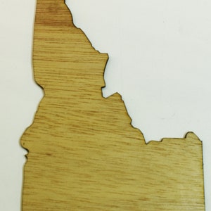 May include: Wooden cutout in the shape of the state of Idaho. The wood has a light brown color with visible grain. The edges are cleanly cut, and the overall shape is well-defined.
