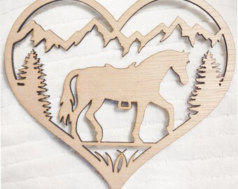 May include: Wooden heart-shaped wall art featuring a horse silhouette, mountains, and trees. The design is cut out of light-colored wood, creating a layered effect. The horse is depicted with a saddle, and the mountains are jagged.