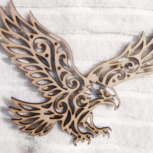 May include: A laser-cut wooden eagle wall decoration featuring intricate scrollwork and cutout patterns. The eagle is depicted in flight with wings spread wide, showcasing a natural light wood finish with dark, charred edges from the cutting process.