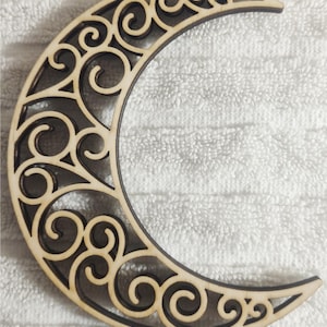 May include: A wooden crescent moon with an intricate, swirling cutout design. The moon is light brown with dark brown edges, creating a contrast that highlights the detailed patterns. The celestial-themed decor is suitable for wall art or craft projects.