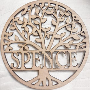 May include: A round, wooden tree of life design with the name "SPENCE" cut out in the center. The tree features detailed branches and leaves, all in a natural wood tone. The design is set against a white background.