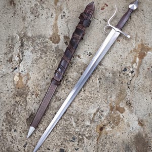 May include: A medieval sword with a polished steel blade and a brown leather-wrapped handle. The sword is accompanied by a matching brown leather sheath. The hilt features a crossguard and a rounded pommel. The overall length of the sword is approximately 36 inches.
