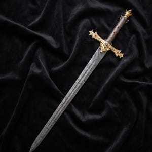 May include: An ornate sword with a patterned blade and gold accents. The hilt features a gold crossguard with red jewels and a detailed design. The sword rests on a black velvet surface, showcasing its intricate craftsmanship.