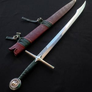May include: A silver sword with a double-edged blade and a cross-shaped hilt, next to a brown leather scabbard with green accents. The sword's handle is wrapped in green leather, and the scabbard has a decorative embossed pattern.