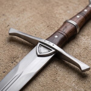 May include: Close-up of a medieval sword with a polished steel blade and a wooden handle. The handle is dark brown with silver accents and a decorative shield near the blade. The sword is resting on a textured, tan surface.