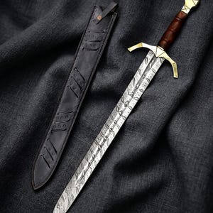 May include: A decorative sword with a patterned blade, a brown wooden handle, and gold-colored crossguard and pommel. The sword is displayed with a black leather sheath. The blade has a detailed, textured design.