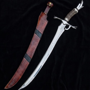 May include: A curved sword with a polished steel blade and a decorative antler handle. The sword is displayed next to a brown leather sheath with black accents. The blade has a curved design and is pointed at the end.