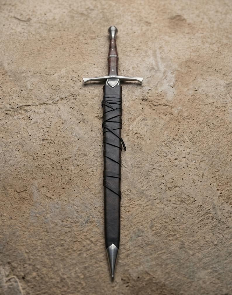 May include: A medieval sword with a silver crossguard and a brown handle. The sword is sheathed in a black leather scabbard, wrapped with leather strips. The tip of the scabbard is silver.