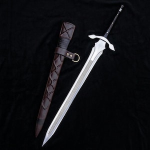 May include: A silver-coloured metal sword with a long, pointed blade featuring a central grooved section, a cross-shaped guard, and a black handle. Beside it lies a dark brown leather scabbard with a decorative embossed pattern and a brass ring.