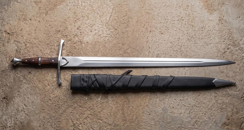 May include: A medieval sword with a polished steel blade and a wooden handle. The sword is laid out next to its black leather sheath. The handle is a dark brown color, and the crossguard is silver.