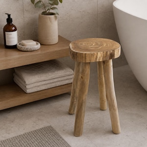May include: A natural wood stool with three legs. The round seat displays the wood's grain. The stool is in a bathroom setting, near a wooden shelf with towels, a soap dish, and a bottle of lotion. The color palette is neutral with beige and brown tones.