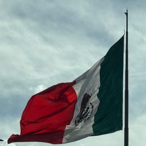 Puede incluir: Una bandera mexicana ondeando al viento contra un cielo nublado. La bandera presenta franjas verticales verdes, blancas y rojas, con el escudo de armas mexicano en el centro de la franja blanca. La bandera está sujeta a un mástil alto y oscuro.