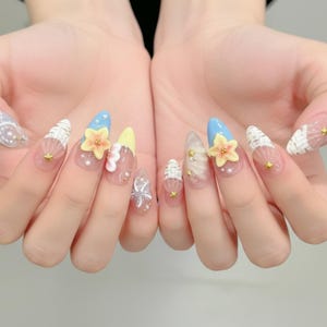 Könnte beinhalten: Nahaufnahme von langen, mandelförmigen Acrylnägeln. Die Nägel weisen verschiedene Designs auf, darunter hellblaue Spitzen, gelbe und orangefarbene Blumenakzente, weiße Muschelmuster und kleine goldene Sternverzierungen. Einige Nägel haben klare Basen mit 3D-Blumendesigns.