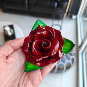 May include: A handcrafted, glossy, deep red rose with green leaves. The rose is held in a hand, showcasing its intricate details and vibrant colours. This decorative piece is likely a brooch or a similar accessory.