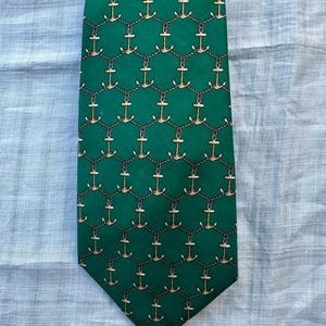 May include: A green necktie featuring a repeating pattern of gold-colored anchors interconnected by dark brown rope-like lines. The design is arranged in a consistent grid across the entire surface of the fabric, creating a nautical-themed motif.