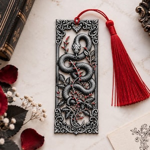 Peut inclure: Un marque-page rectangulaire en métal orné d'un relief détaillé représentant un serpent enroulé parmi des branches feuillues. Le motif est fini dans un ton argenté sombre avec des touches rouges sur les feuilles et les yeux du serpent.
