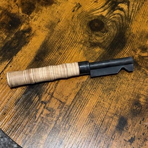May include: A black and tan-colored object with a wooden handle and a metal end. The handle is light brown with a textured, wrapped design. The metal end is black and has a notched design. The object is lying on a wooden surface.