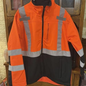 May include: A high-visibility jacket featuring a bright orange upper section and a black lower section. The garment includes vertical and horizontal silver reflective strips on the chest, shoulders, and sleeves, a full-length black front zipper, and a chest pocket.