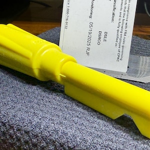 May include: A bright yellow plastic tool with a thick, cylindrical handle and a flat, extended shaft featuring a notched end. The tool is positioned horizontally, showcasing its molded construction and uniform, vibrant yellow color throughout.