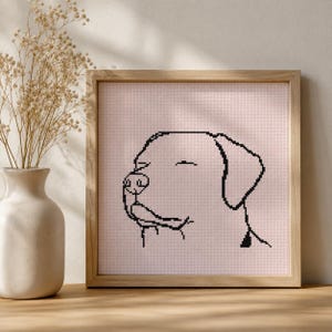 May include: A square wooden frame containing a cross-stitch embroidery of a dog profile. The design features a black stitched outline of a dog head on a light pink fabric background. The dog is depicted with closed eyes and a simple snout shape.
