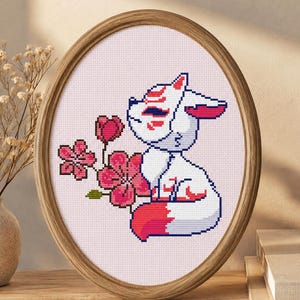 May include: An oval wooden frame containing a cross-stitch embroidery of a white fox wearing a red and white patterned mask. The fox is positioned next to a cluster of pink cherry blossom flowers against a light pink grid background.