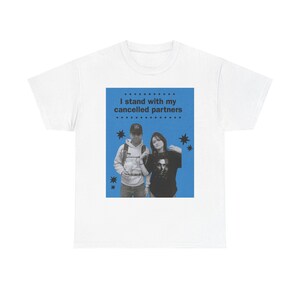 Può includere: T-shirt bianca con un quadrato blu con due persone e la scritta "I stand with my cancelled partners". Il design include grafiche a stella. La maglietta è un capo casual.